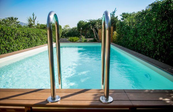 Piscina con scaletta in metallo, acqua turchese e terrazza in legno circondata da cespugli verdi sotto un cielo azzurro.