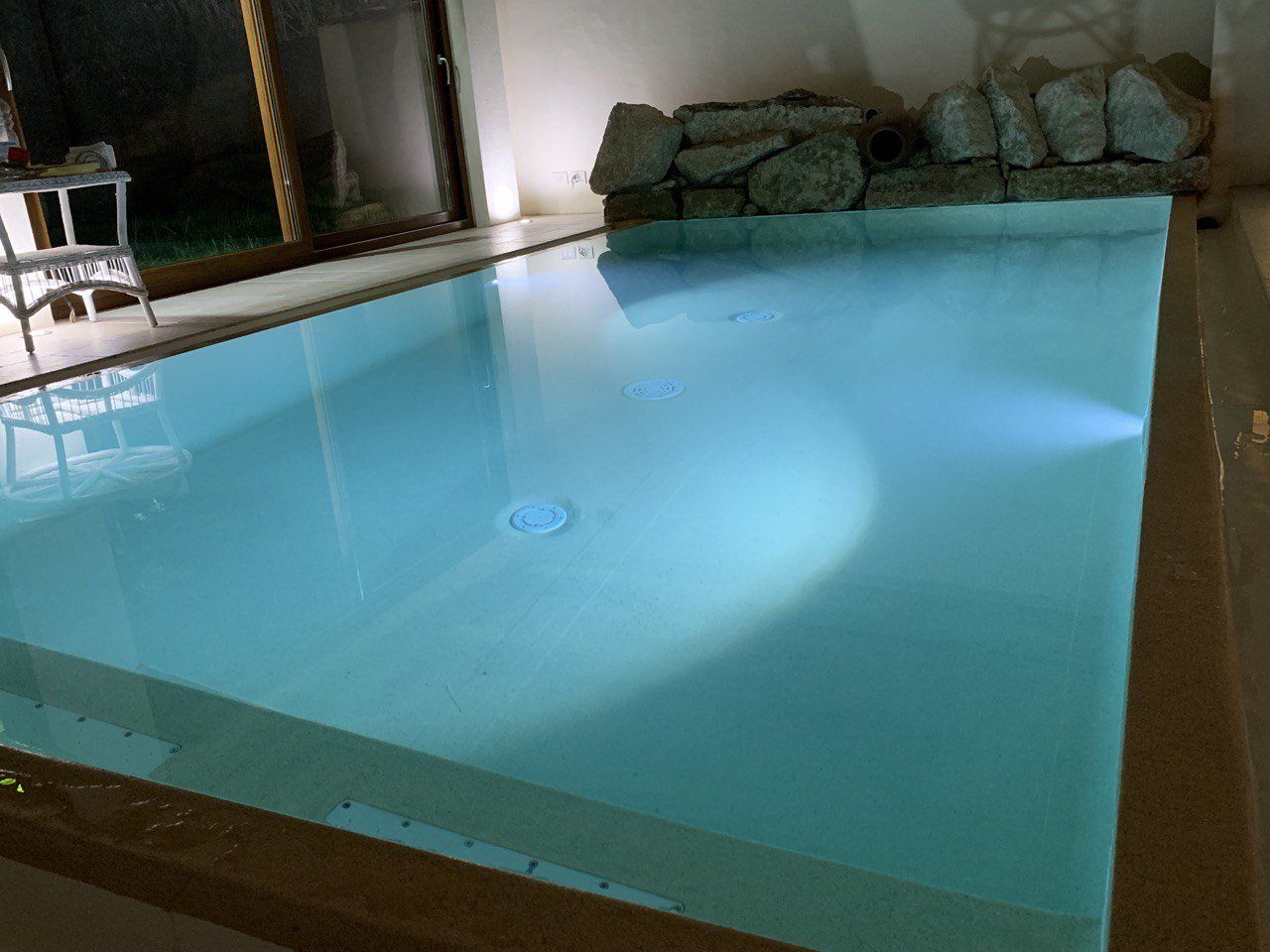 Piscina interna rettangolare con acqua blu, illuminata da faretti. Rocce decorative sullo sfondo.
