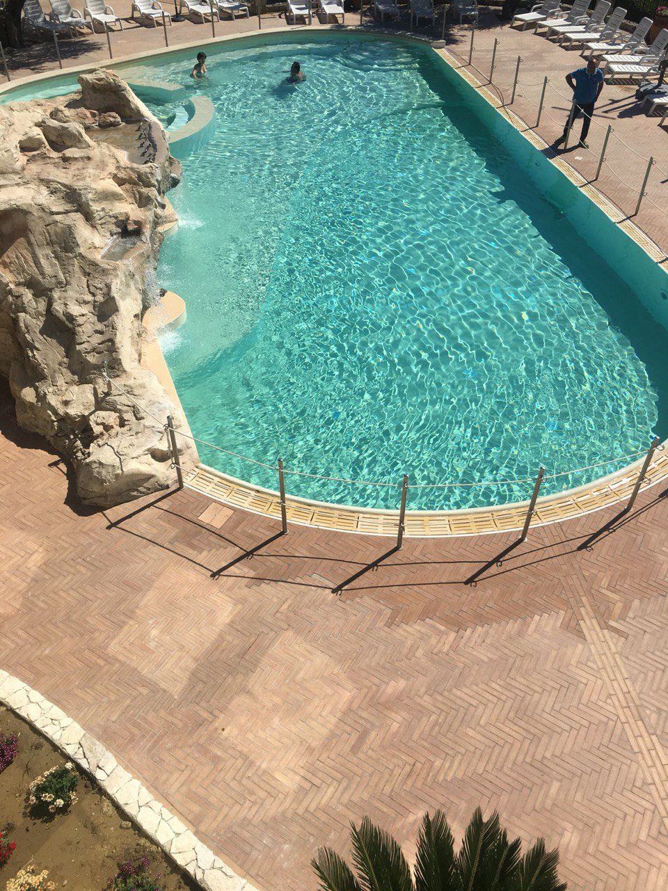 Piscina con persone che nuotano, circondata da un patio in mattoni e sedie a sdraio.