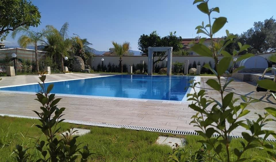 Piscina con acqua azzurra circondata da un patio in pietra, erba verde e alberi sotto un cielo limpido.