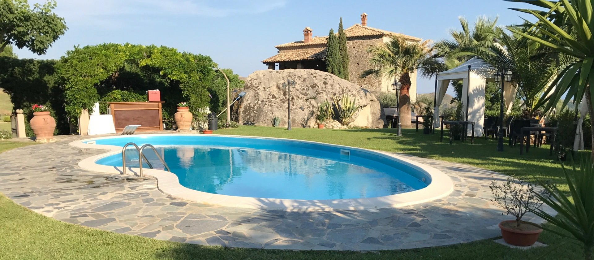Una piscina blu in un lussureggiante giardino verde, con una casa in pietra sullo sfondo.