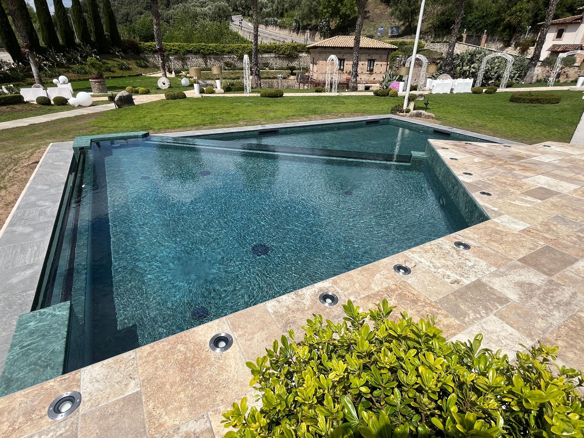 Piscina con acqua scura e patio in pietra, circondata da prato verde e giardino.