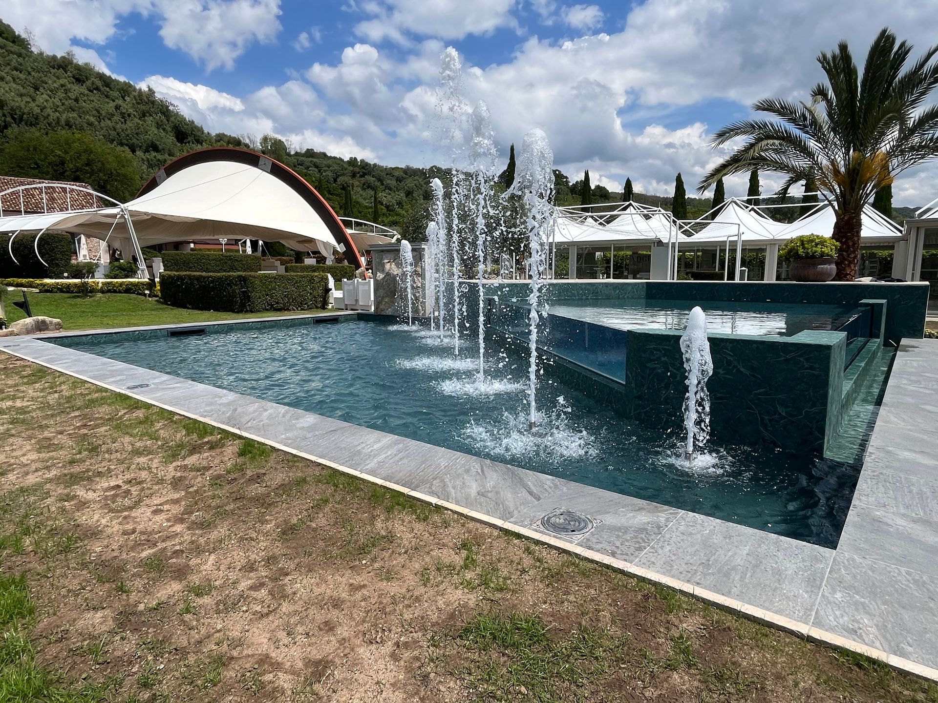 La fontana spruzza l'acqua in una piscina circondata da giardini, capanne e un ambiente esterno soleggiato e collinare.