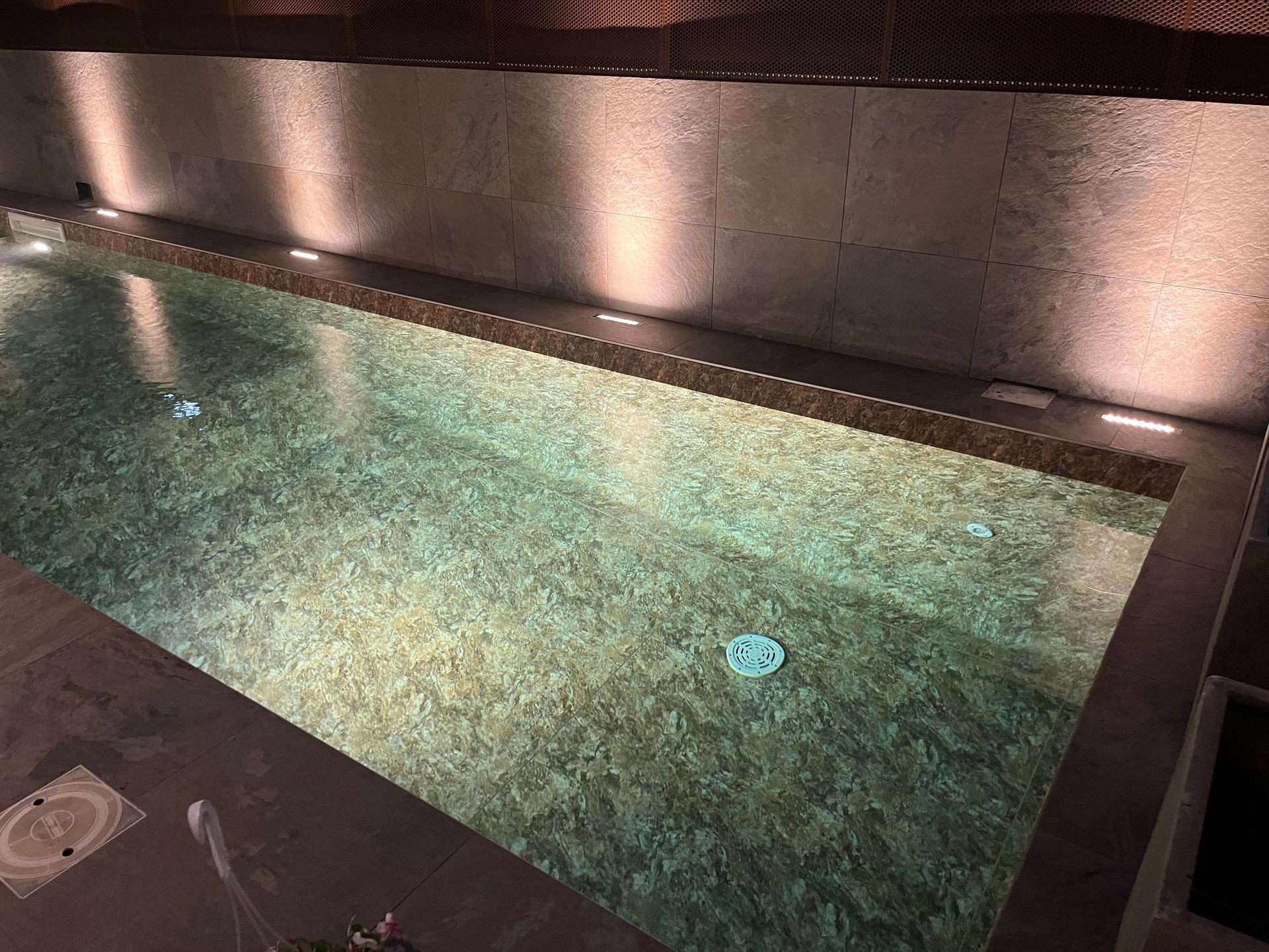 Piscina coperta con acqua illuminata, finiture in pietra scura e luci lungo il muro di mattoni.