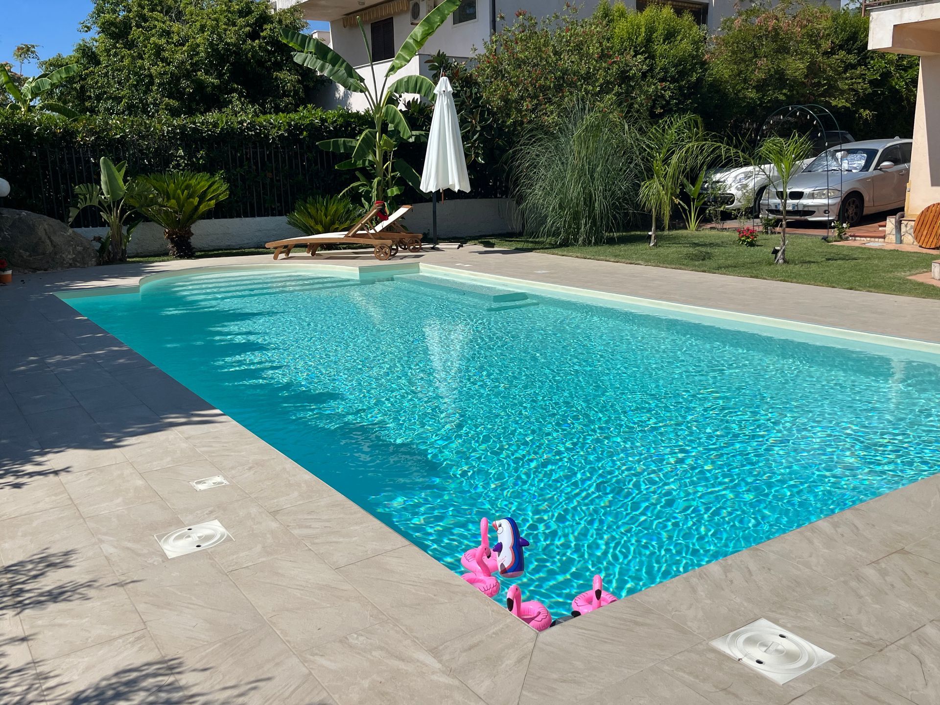 Piscina con acqua turchese, patio, prato ed edifici sotto un cielo soleggiato.