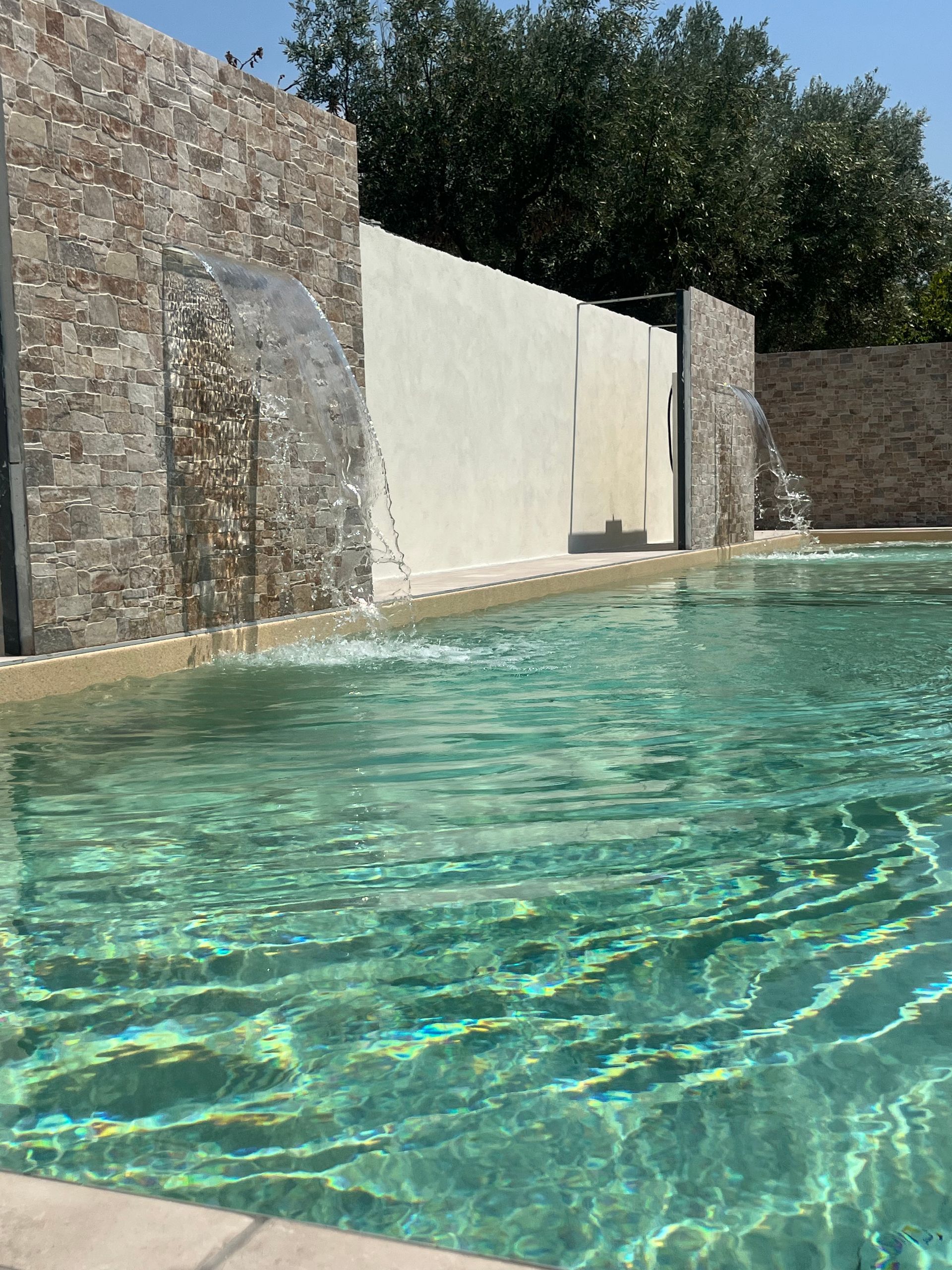 Piscina con acqua limpida, un muro di pietra con una cascata e un muro bianco dietro.
