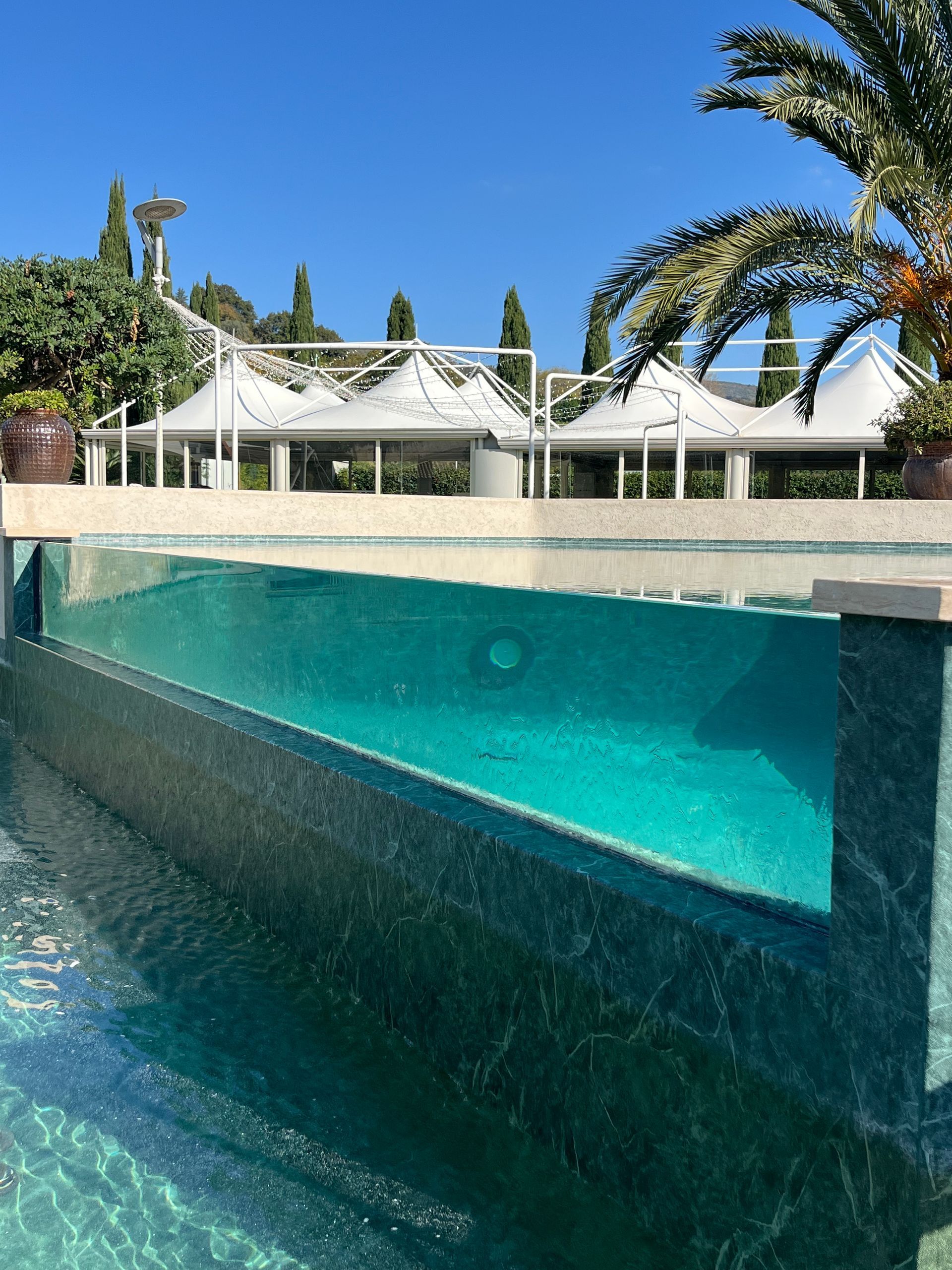 Piscina con pareti in vetro trasparente, acqua turchese, struttura a baldacchino bianco, cielo azzurro.
