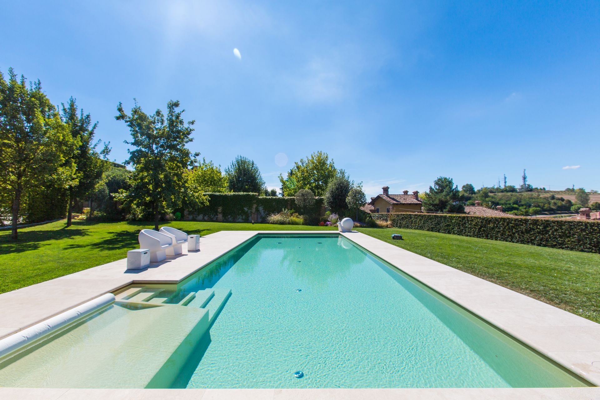 Piscina rettangolare con acqua limpida, circondata da erba verde e alberi, sotto un cielo azzurro.