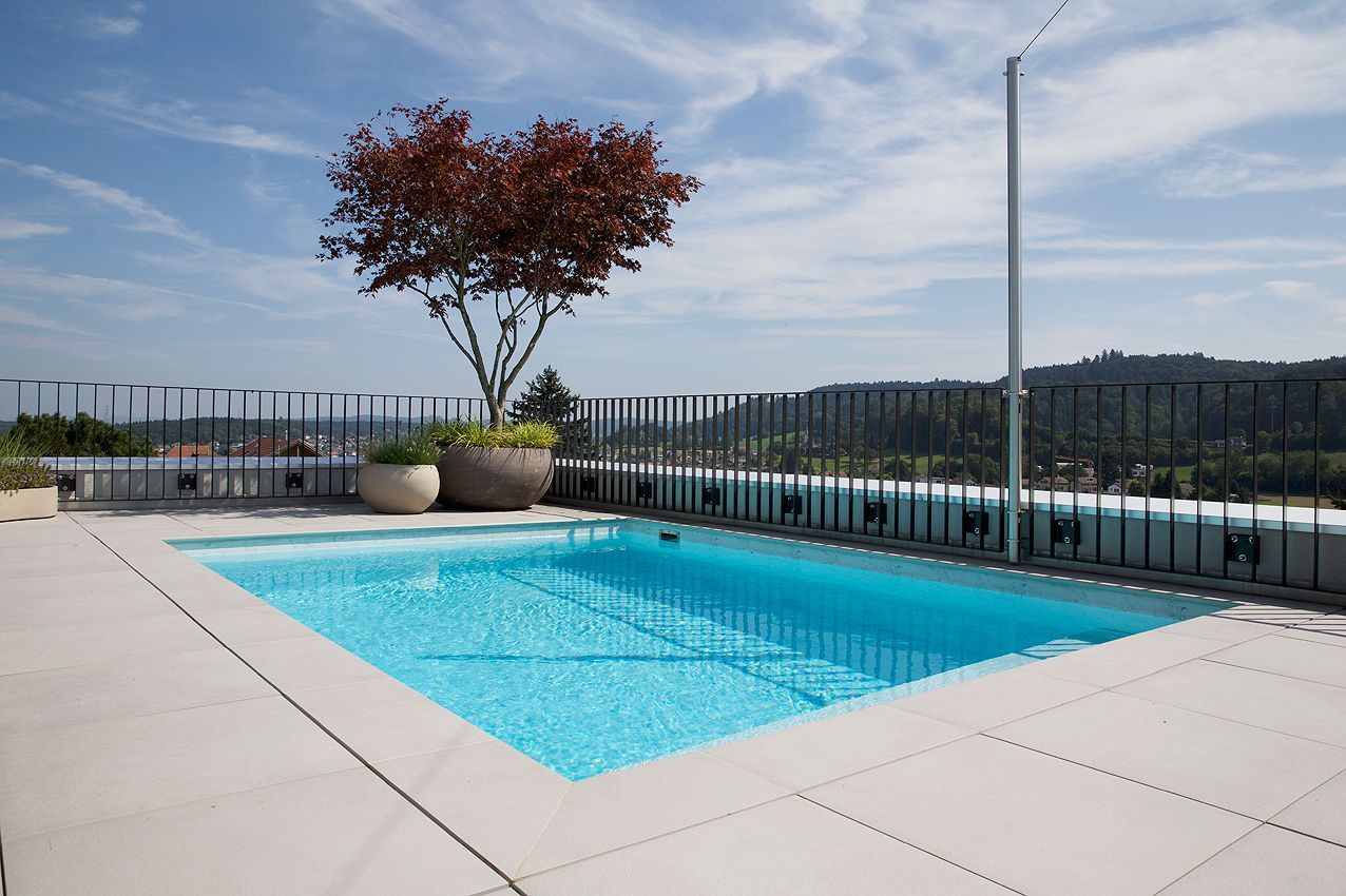 Piscina sul tetto con acqua turchese, albero dalle foglie rosse e vista sulla città sotto un cielo azzurro.