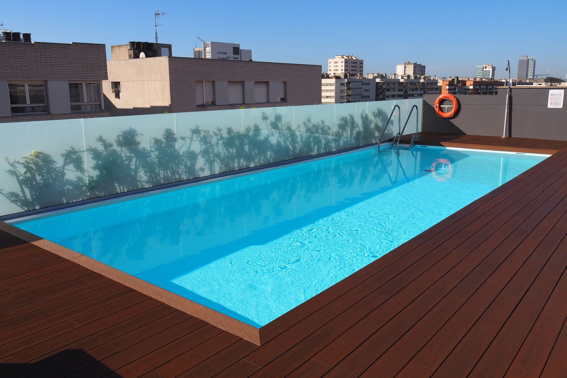 Piscina sul tetto con acqua turchese, terrazza in legno, barriera in vetro e vista sullo skyline della città.