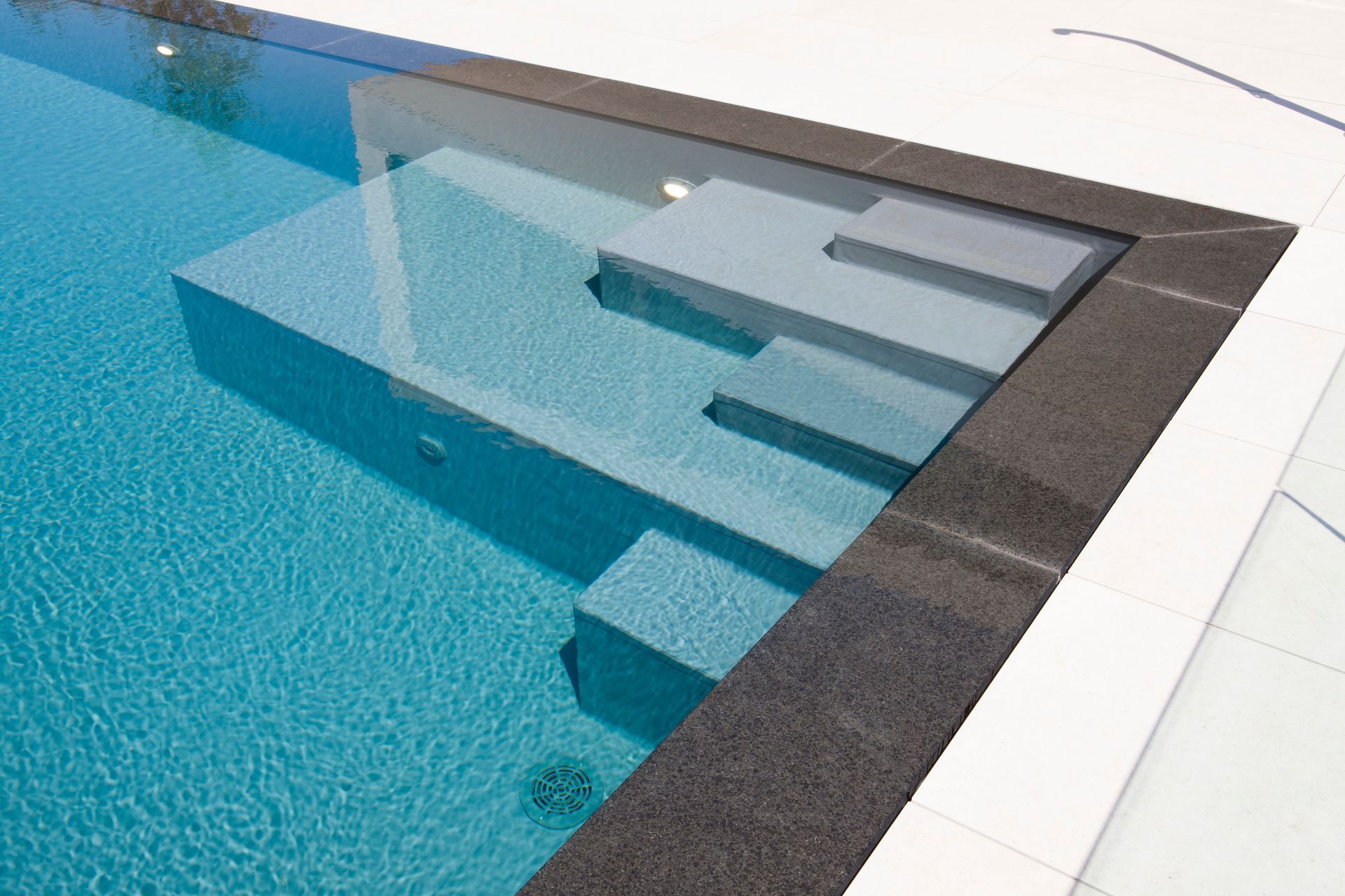 Piscina con gradini integrati, acqua turchese, bordo scuro, terrazza bianca.