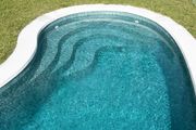 Una piscina con acqua turchese, bordo bianco e gradini, circondata da erba verde.