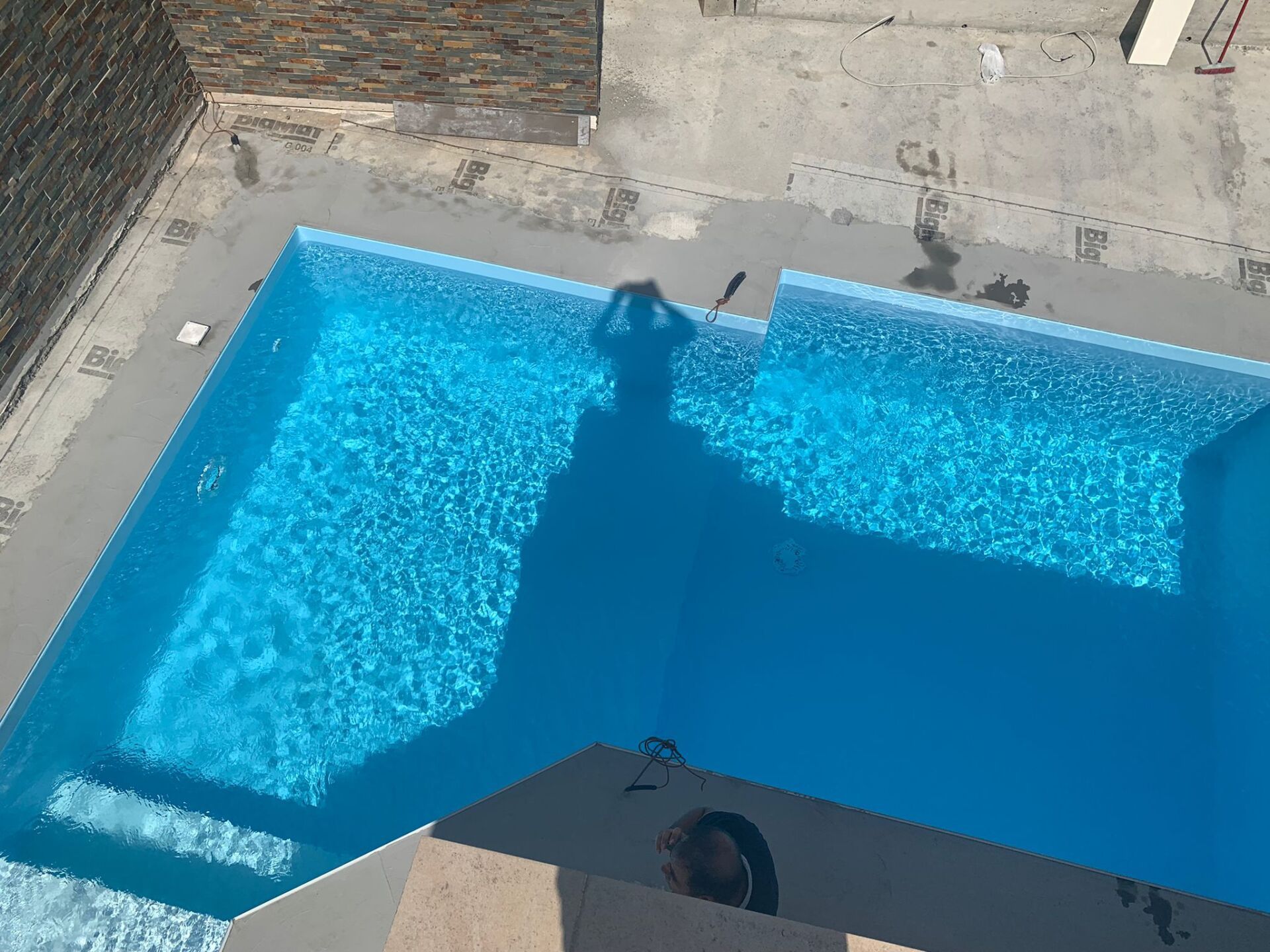 Ripresa dall'alto di una piscina rettangolare blu con increspature visibili. L'ombra di una persona si proietta nell'acqua.