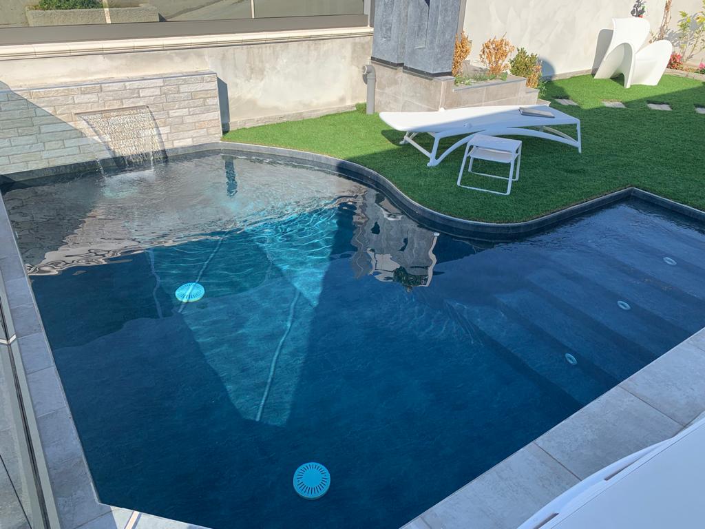 Piscina con cascata, erba artificiale e sedie a sdraio bianche.