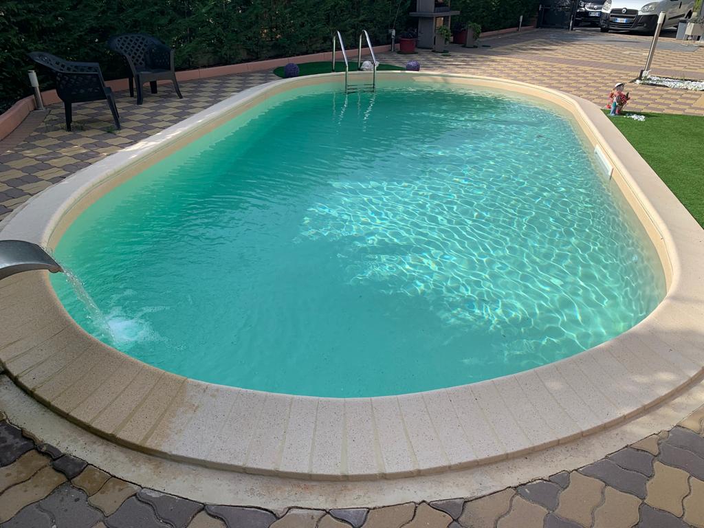 Piscina ovale con acqua limpida, circondata da pareti in pietra chiara. Una fontana spruzza l'acqua nella piscina.