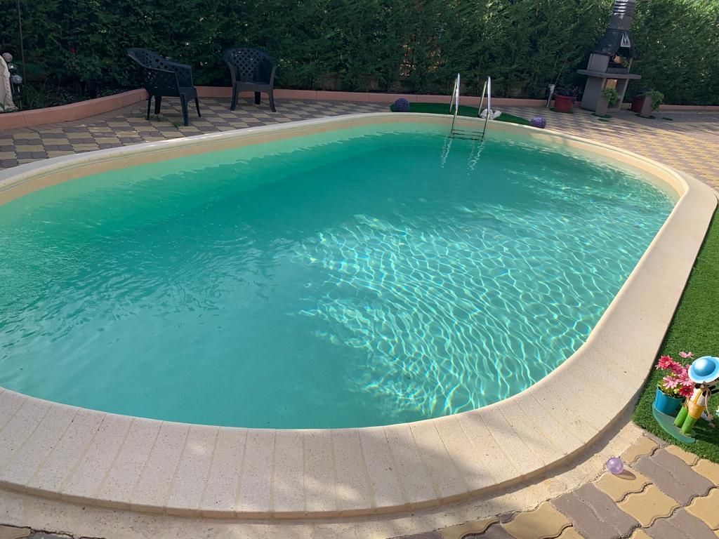 Piscina ovale con acqua turchese e limpida, circondata da cemento color marrone chiaro e da un giardino paesaggistico.