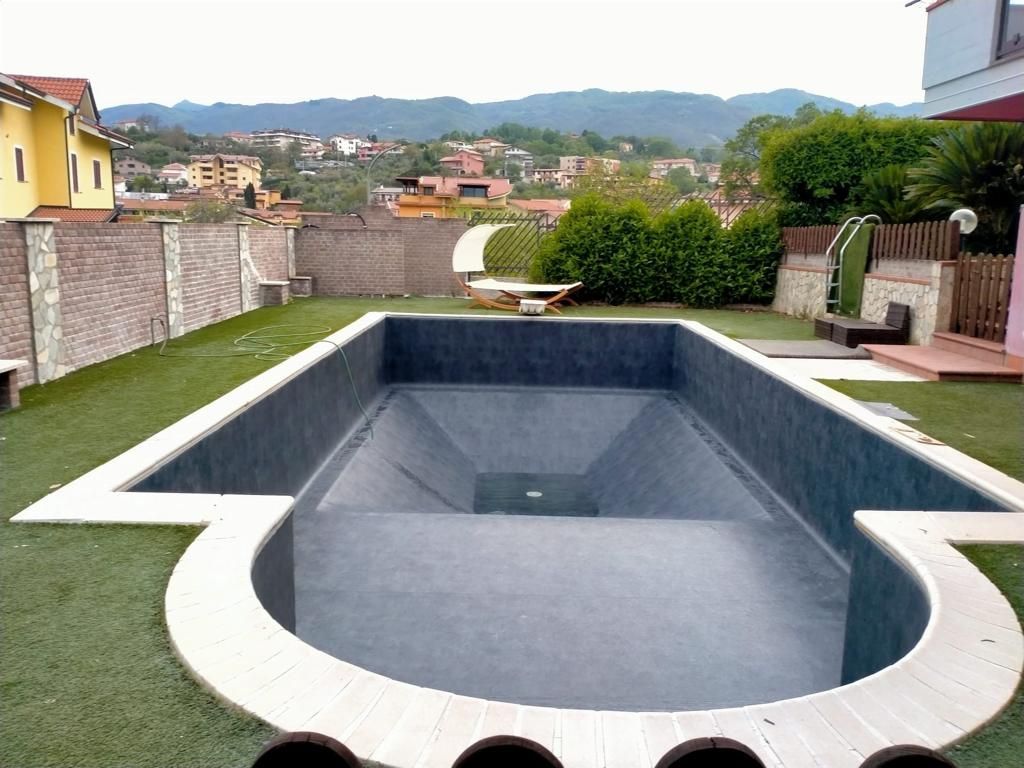 Piscina rettangolare vuota con interno scuro, bordo chiaro e prato in un cortile, con vista sulla città e sulle montagne.