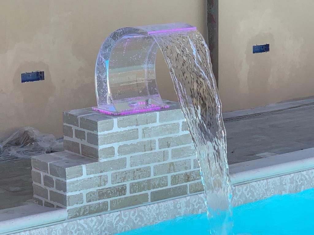 L'acqua cade a cascata da un arco in acrilico illuminato in una piscina. La struttura poggia su mattoni.