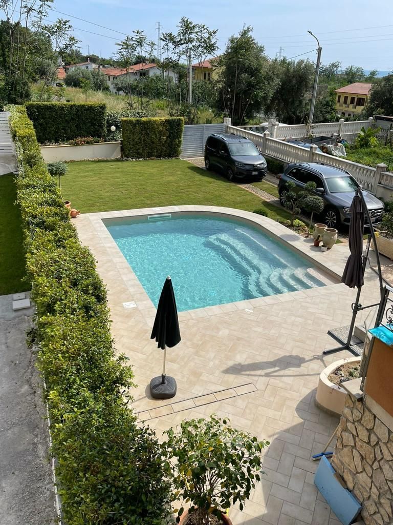 Giardino con piscina, prato e auto parcheggiate, circondato dal verde e dagli edifici, sotto un cielo azzurro.