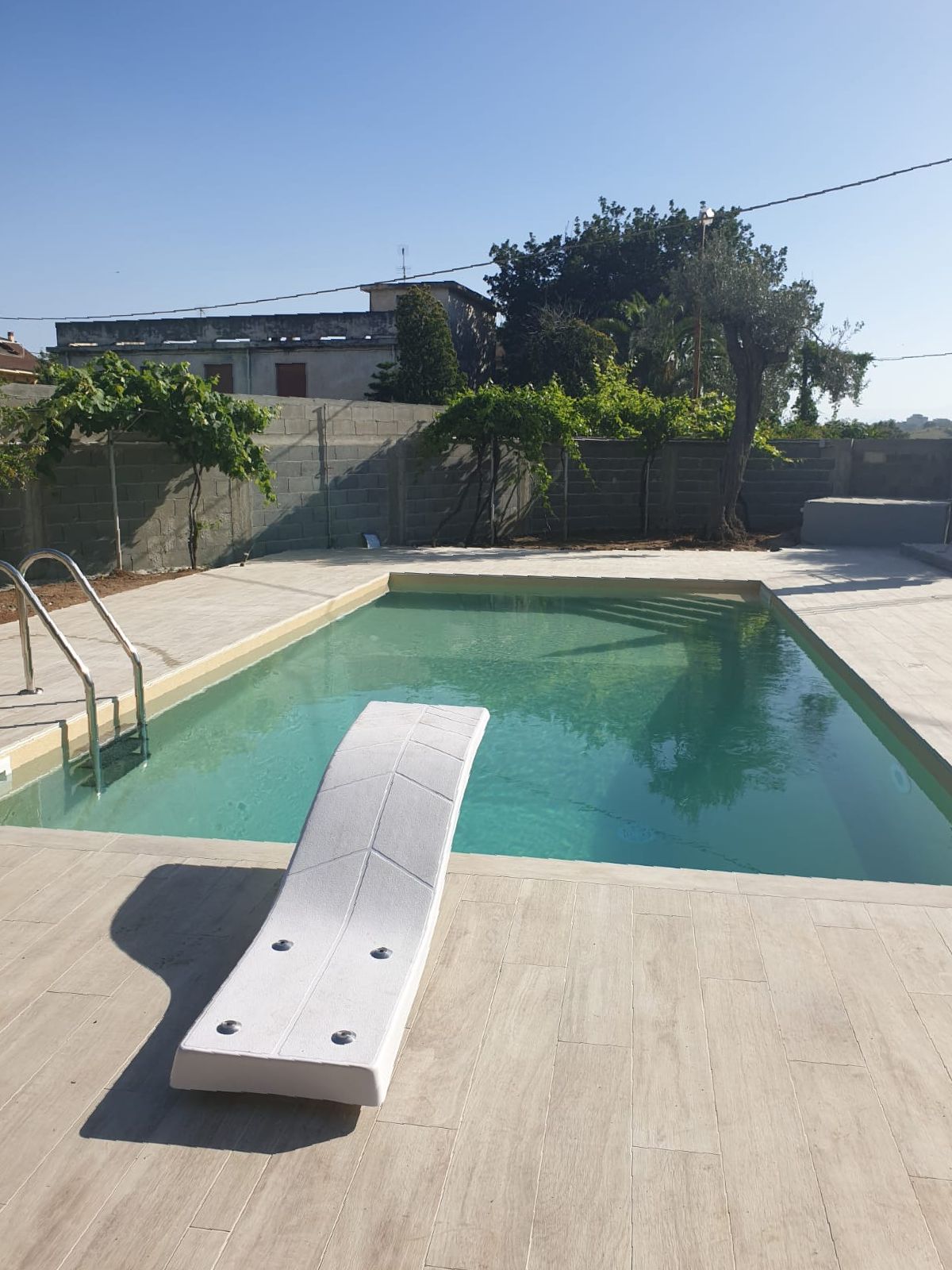 Piscina con trampolino, circondata da cemento, alberi ed edifici. Giornata di sole.