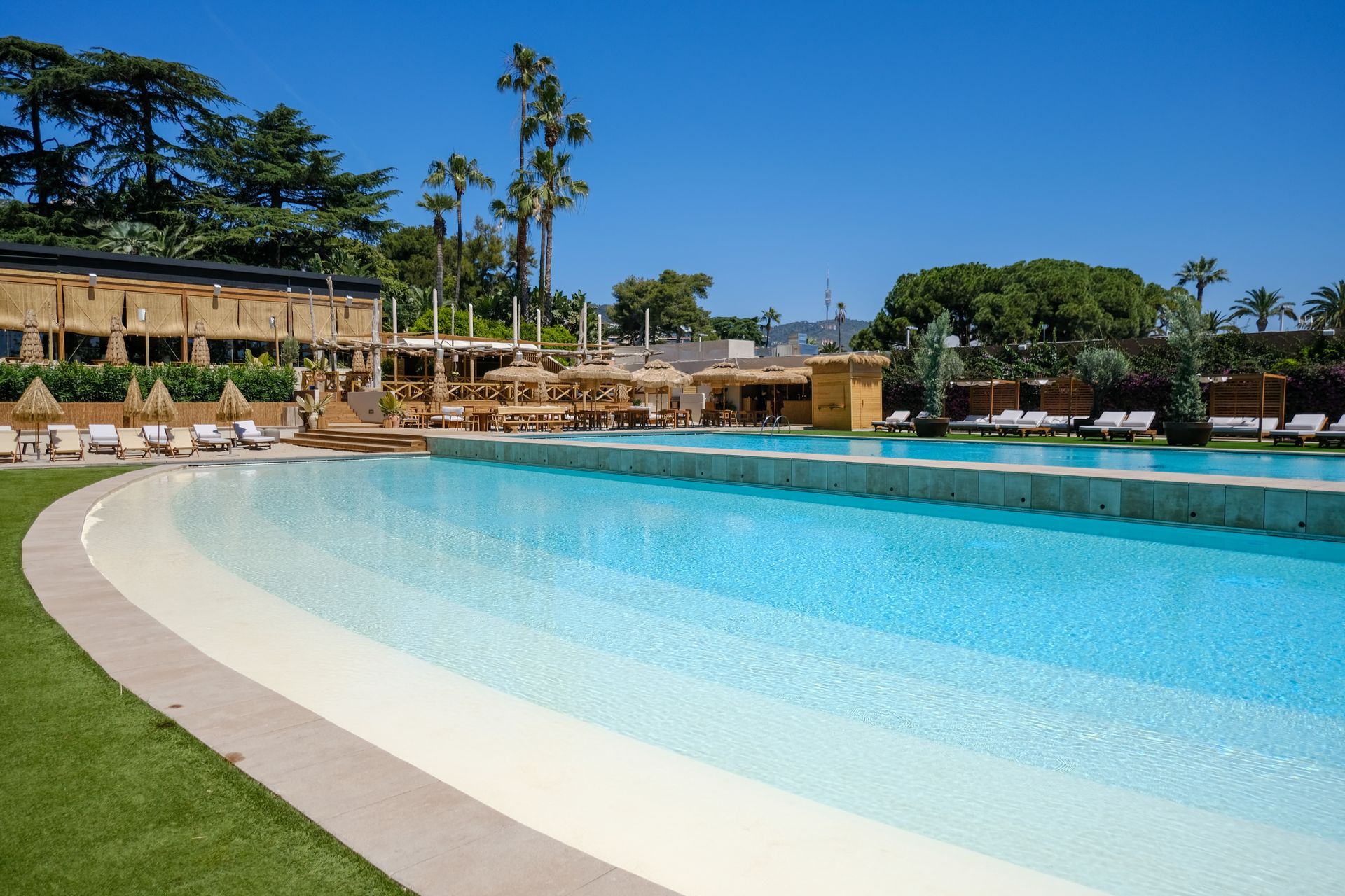 Ampia piscina all'aperto con bordo basso e sabbia bianca. Sedie a sdraio, edifici e palme sullo sfondo. Cielo azzurro.