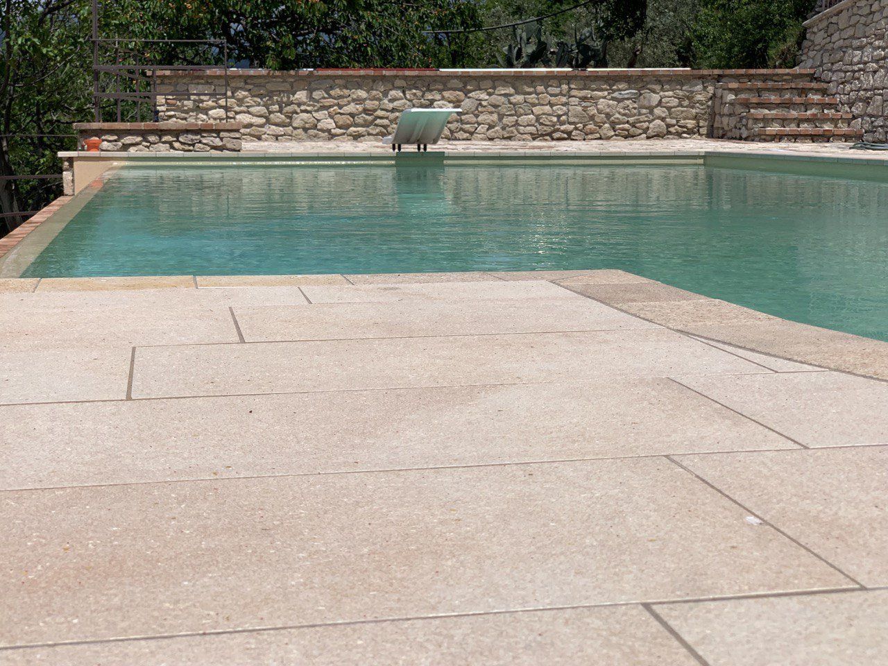 Vista a bordo piscina con patio in pietra che conduce alla piscina trasparente. Muro in pietra e vegetazione sullo sfondo.
