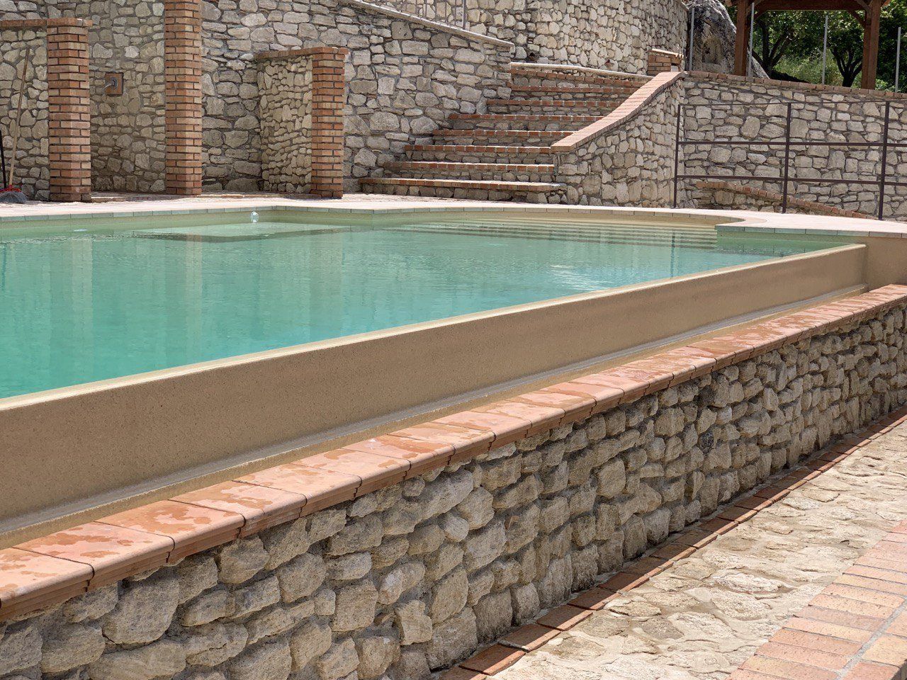 Piscina con pareti in pietra e finiture in mattoni. Una scalinata conduce a una struttura sullo sfondo. Acqua turchese.