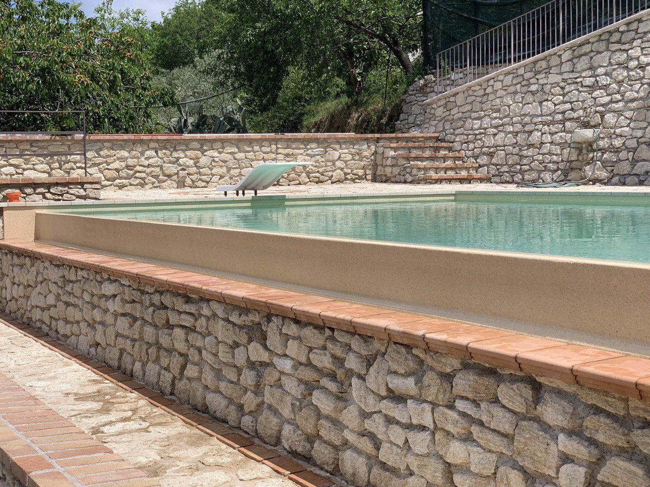 Piscina rettangolare con pareti in pietra e acqua limpida, circondata da pietre e mattoni, con alberi sullo sfondo.