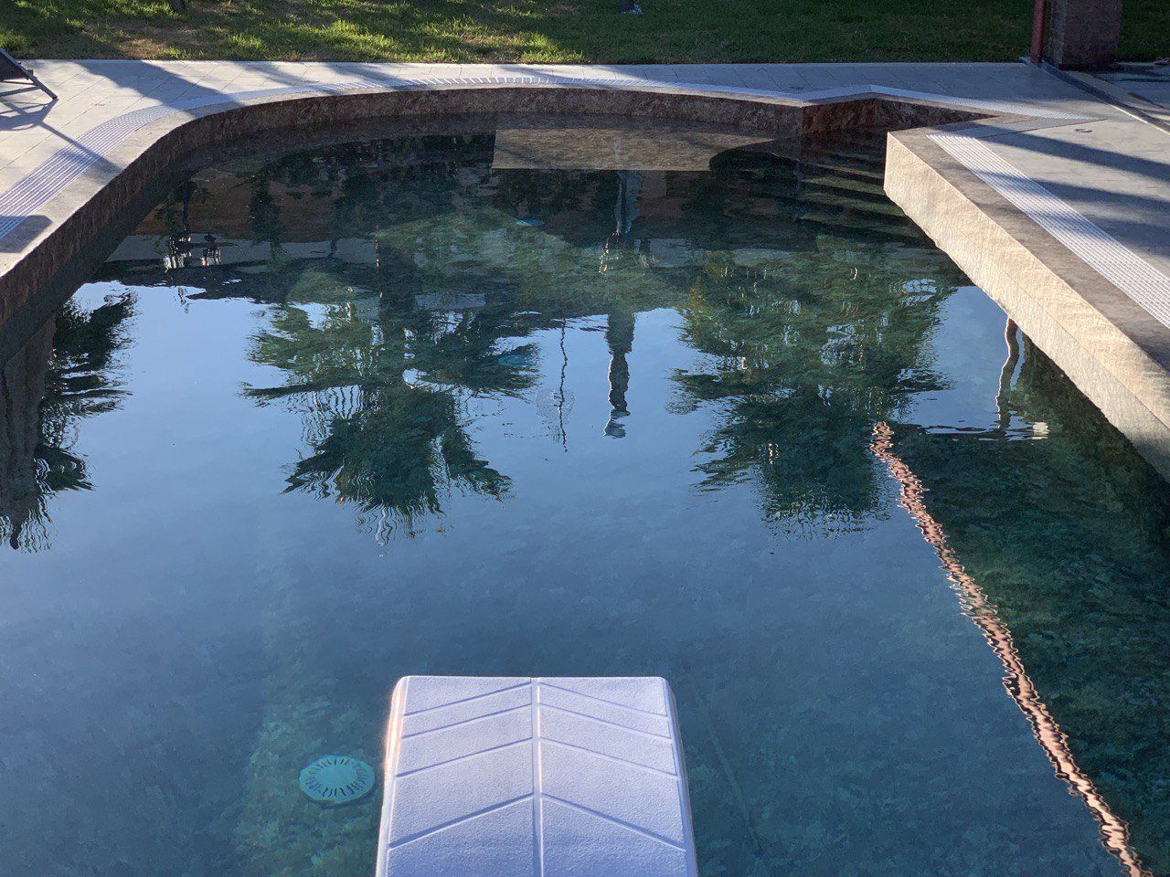 Piscina con trampolino, alberi che si riflettono sotto un cielo soleggiato.