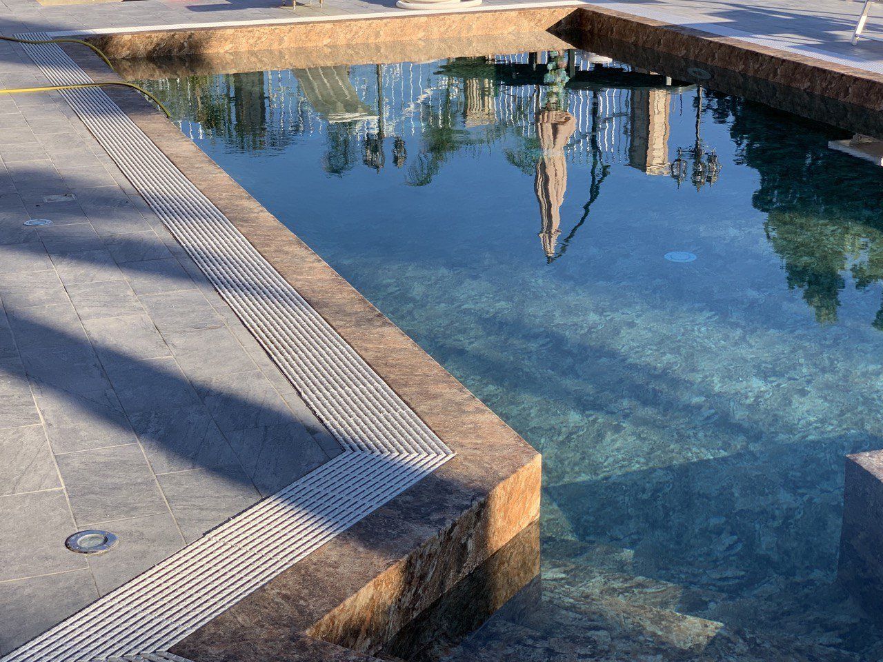 Una piscina riflettente con acqua limpida che riflette un edificio, delimitata da pietre e da uno scarico metallico.