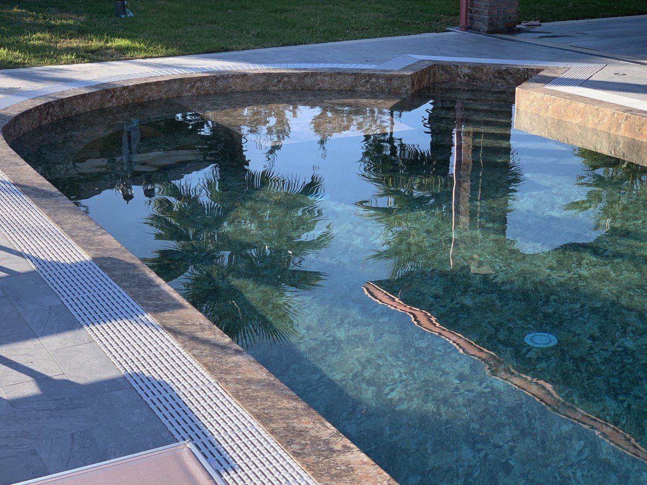 Piscina con riflessi di alberi e cielo. Bordo in pietra e patio circostante.