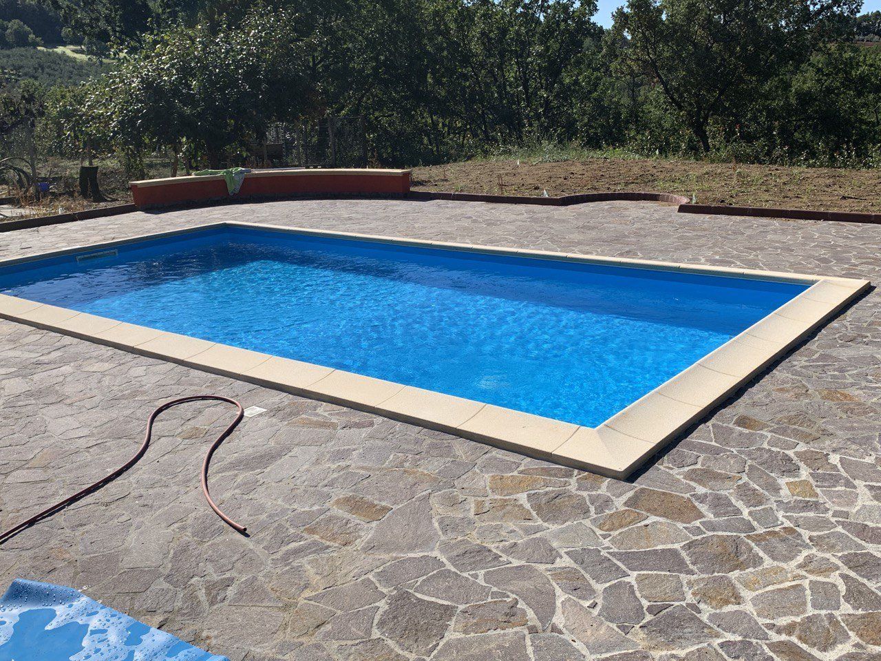 Piscina blu rettangolare circondata da un patio in pietra, con alberi sullo sfondo.
