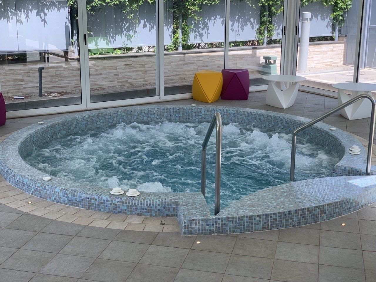 Vasca idromassaggio con piastrelle blu, situata all'interno di una veranda con pareti in vetro e accesso tramite gradini.