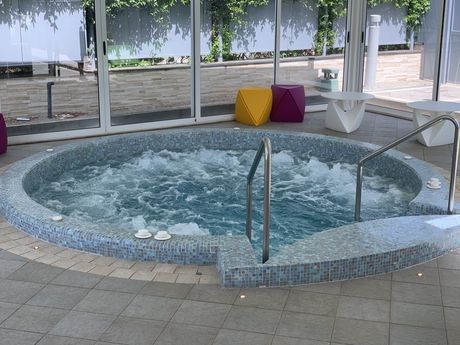 Vasca idromassaggio con piastrelle blu, situata all'interno di una veranda con pareti in vetro e accesso tramite gradini.