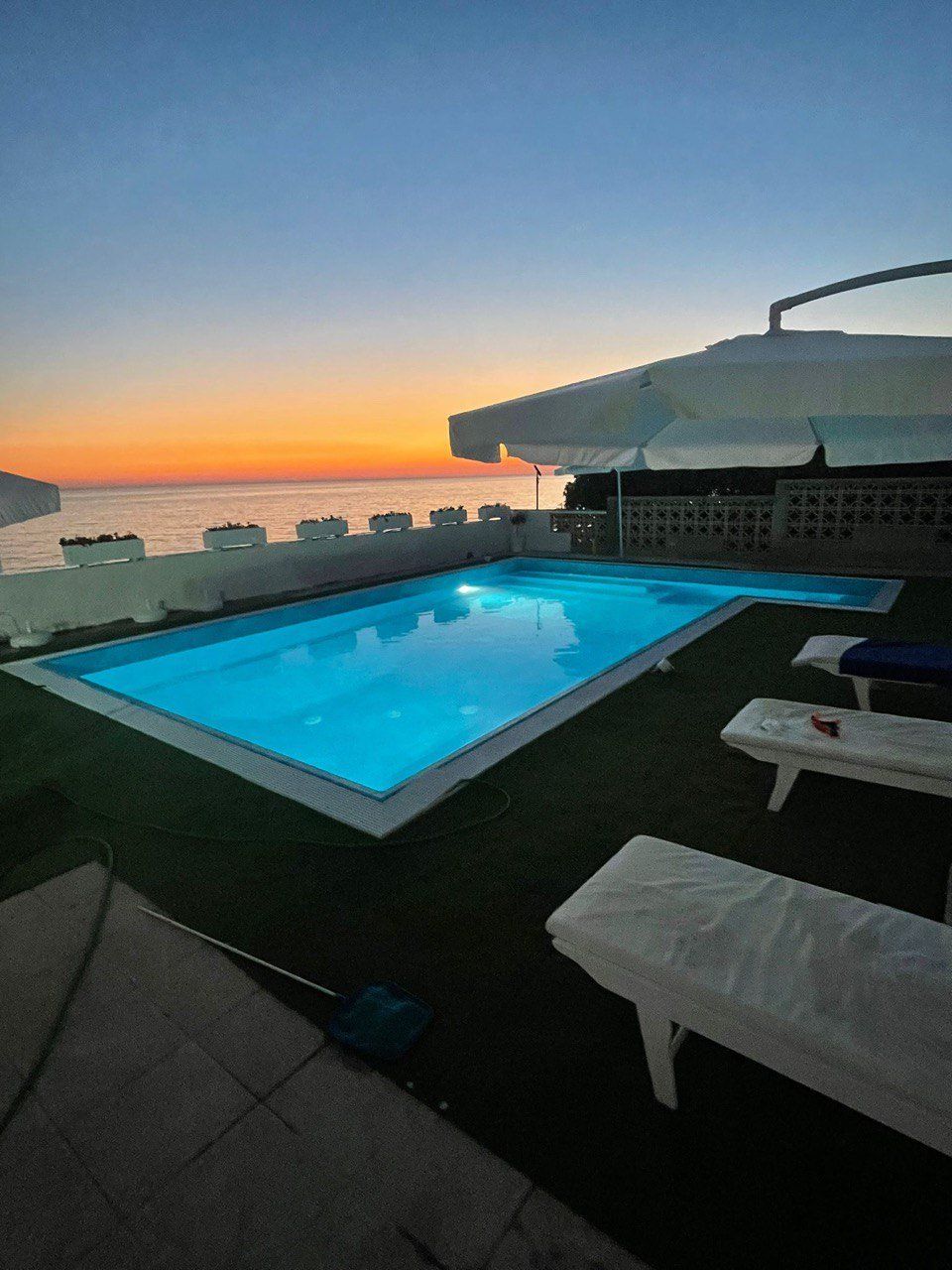 Una piscina rettangolare con acqua azzurra su un tetto al tramonto. Due sedie a sdraio bianche sono sedute lì vicino.