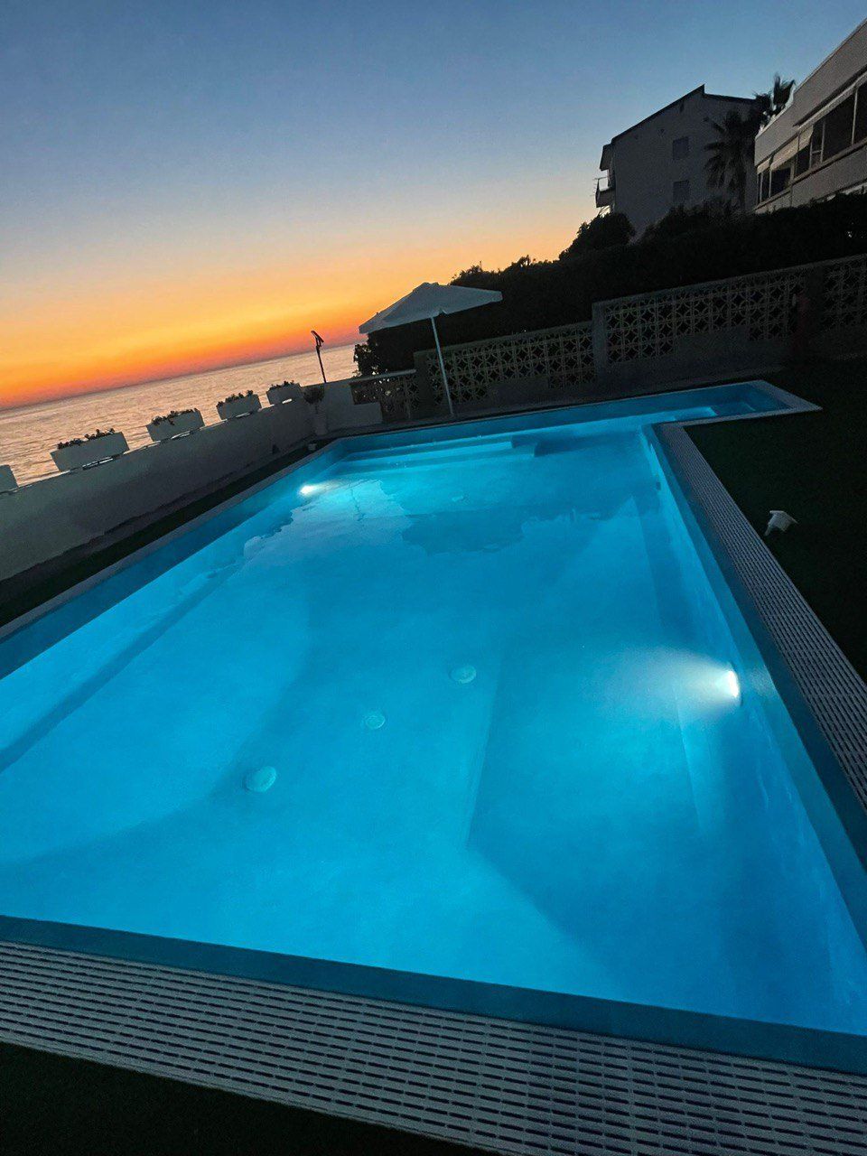 Piscina con vista sull'oceano al tramonto: acqua azzurra, cielo arancione ed edifici bianchi.