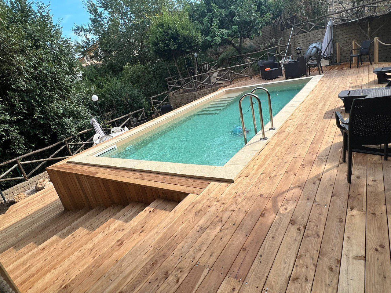 Terrazza in legno con gradini che conducono a una piscina rettangolare; alberi e lucine sullo sfondo.