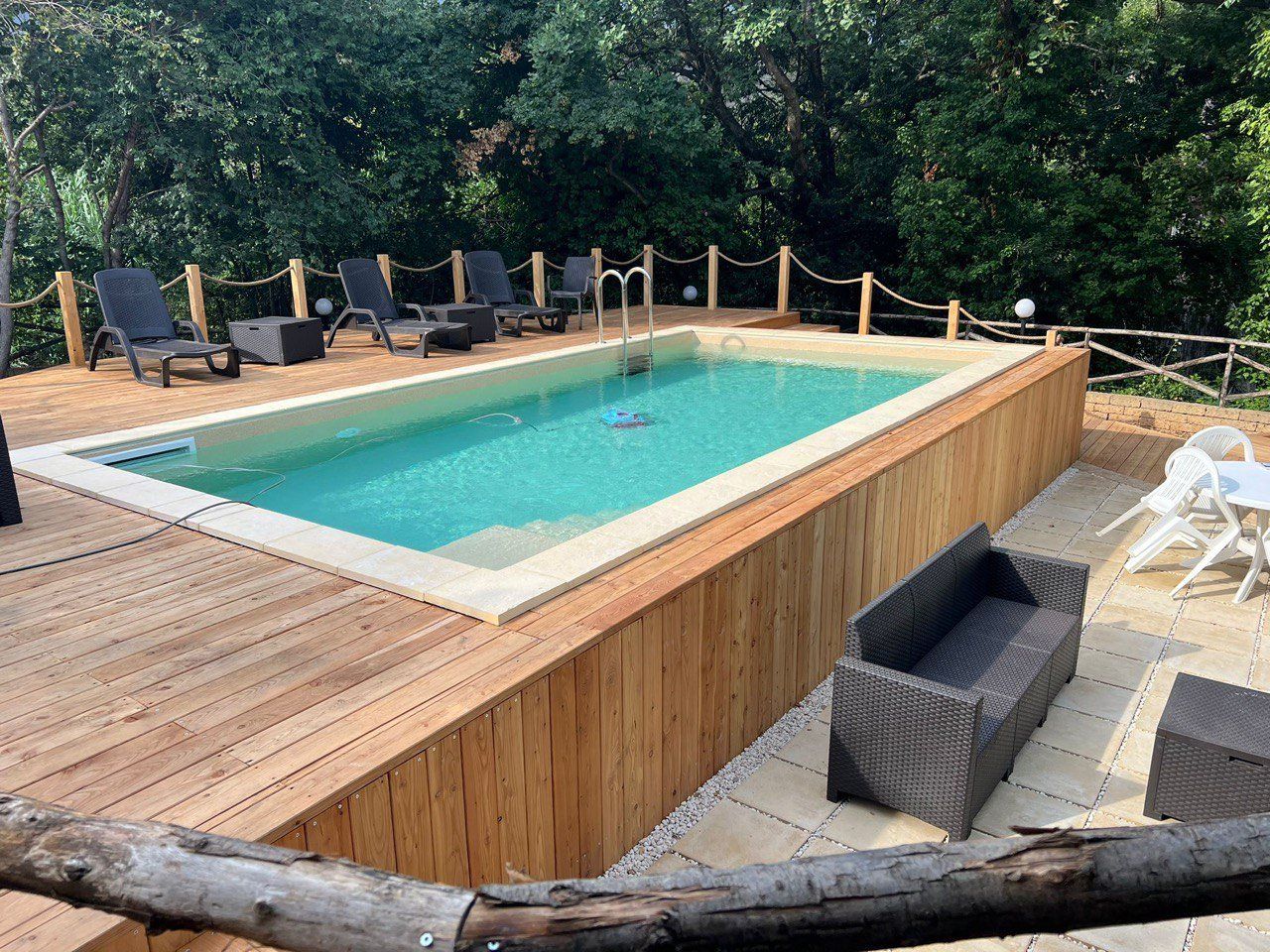 Piscina all'aperto con terrazza in legno, sedie a sdraio e posti a sedere, circondata da alberi.