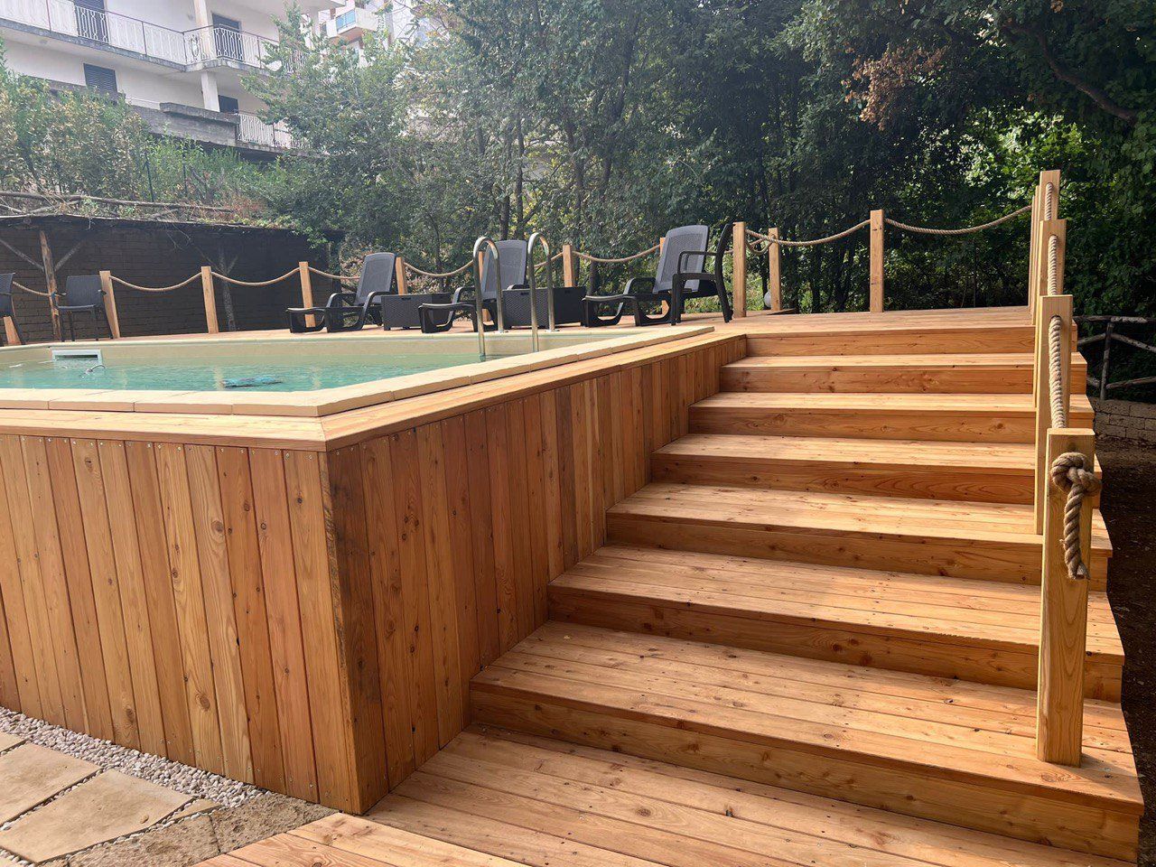 Terrazza in legno e scale che conducono a una piscina con sedie a sdraio, immersa nel verde.