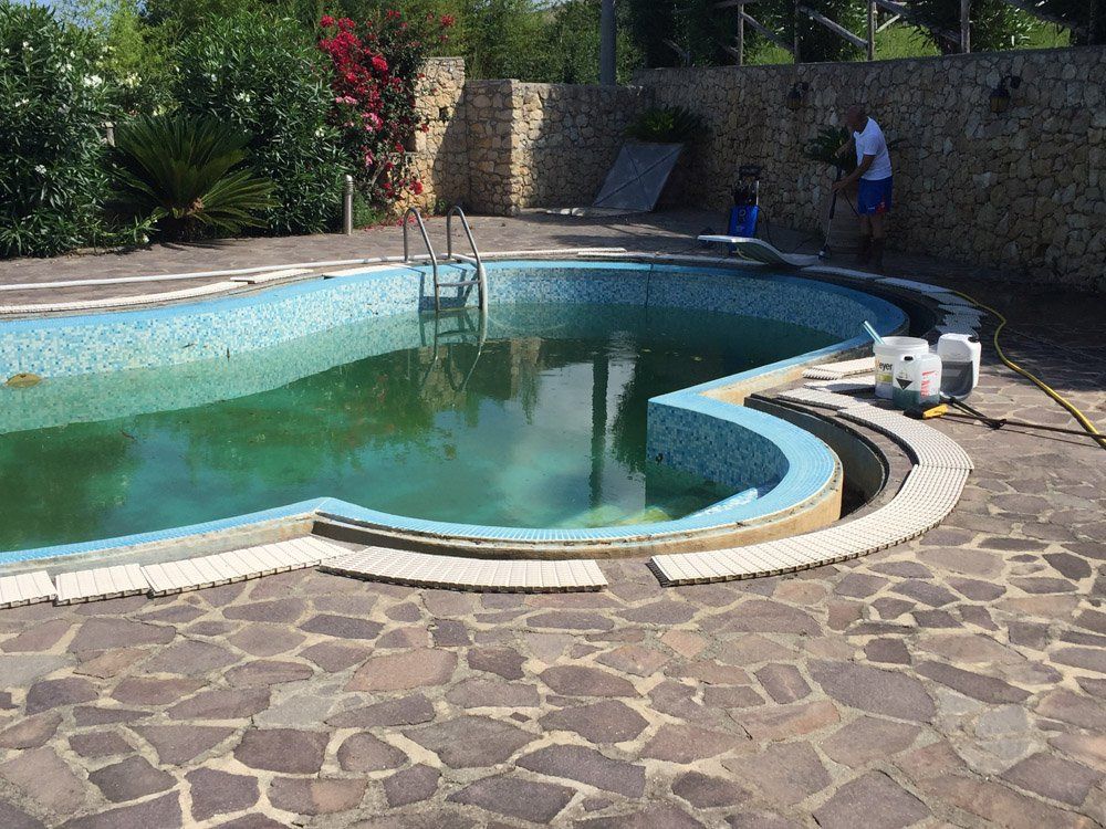 Una piscina sporca, con piastrelle turchesi, è circondata da pietre e da un muro. Un uomo sta lavorando sul muro.