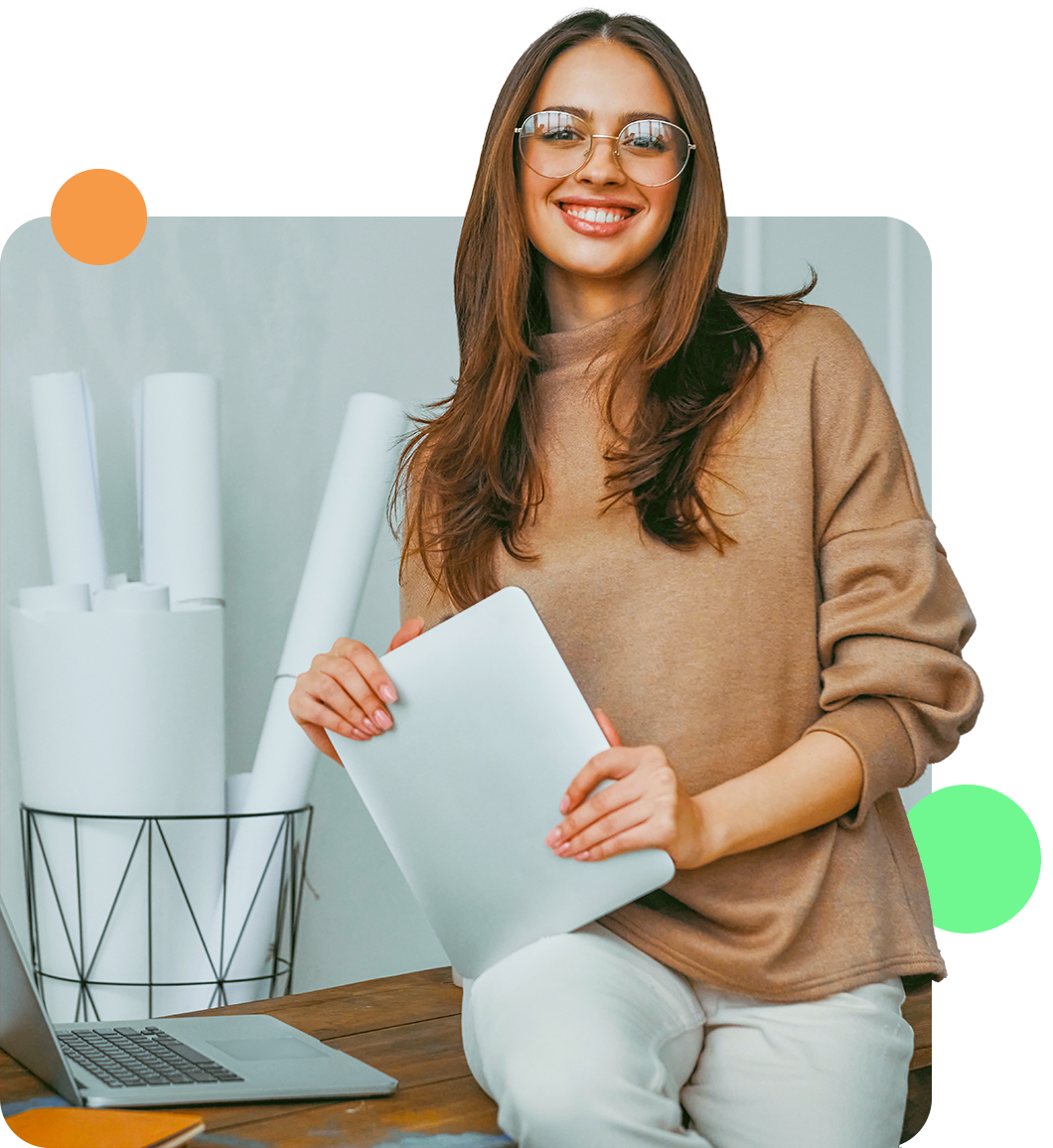 Woman smiling, holding a laptop, sitting by a desk with rolled papers and a laptop in an office.