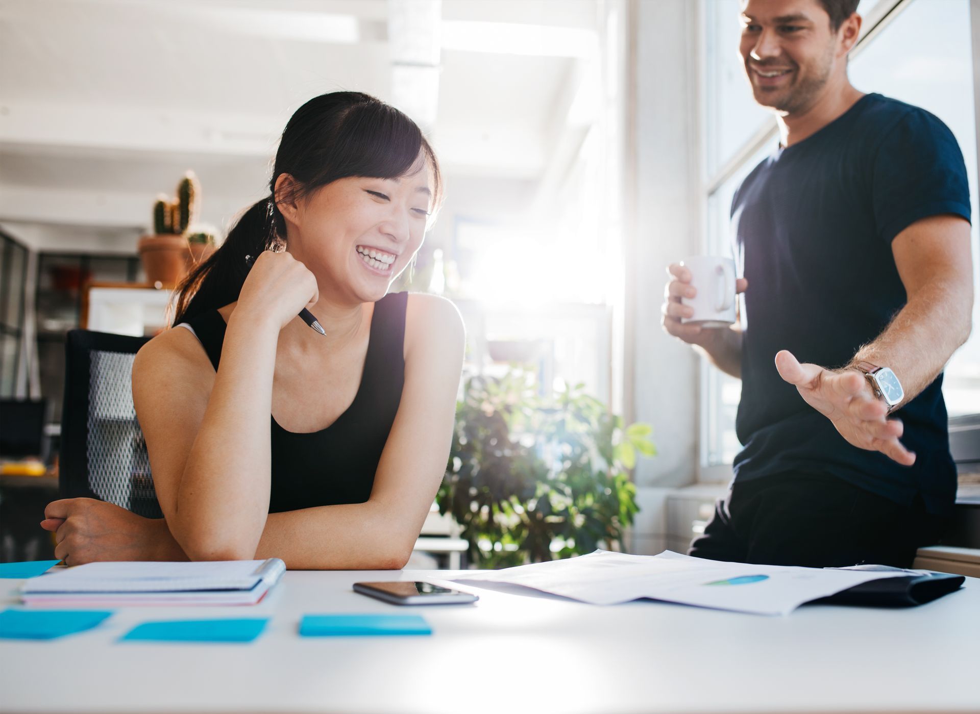 Woman smiles while looking at papers with a man, in an office. Bright sunlight.