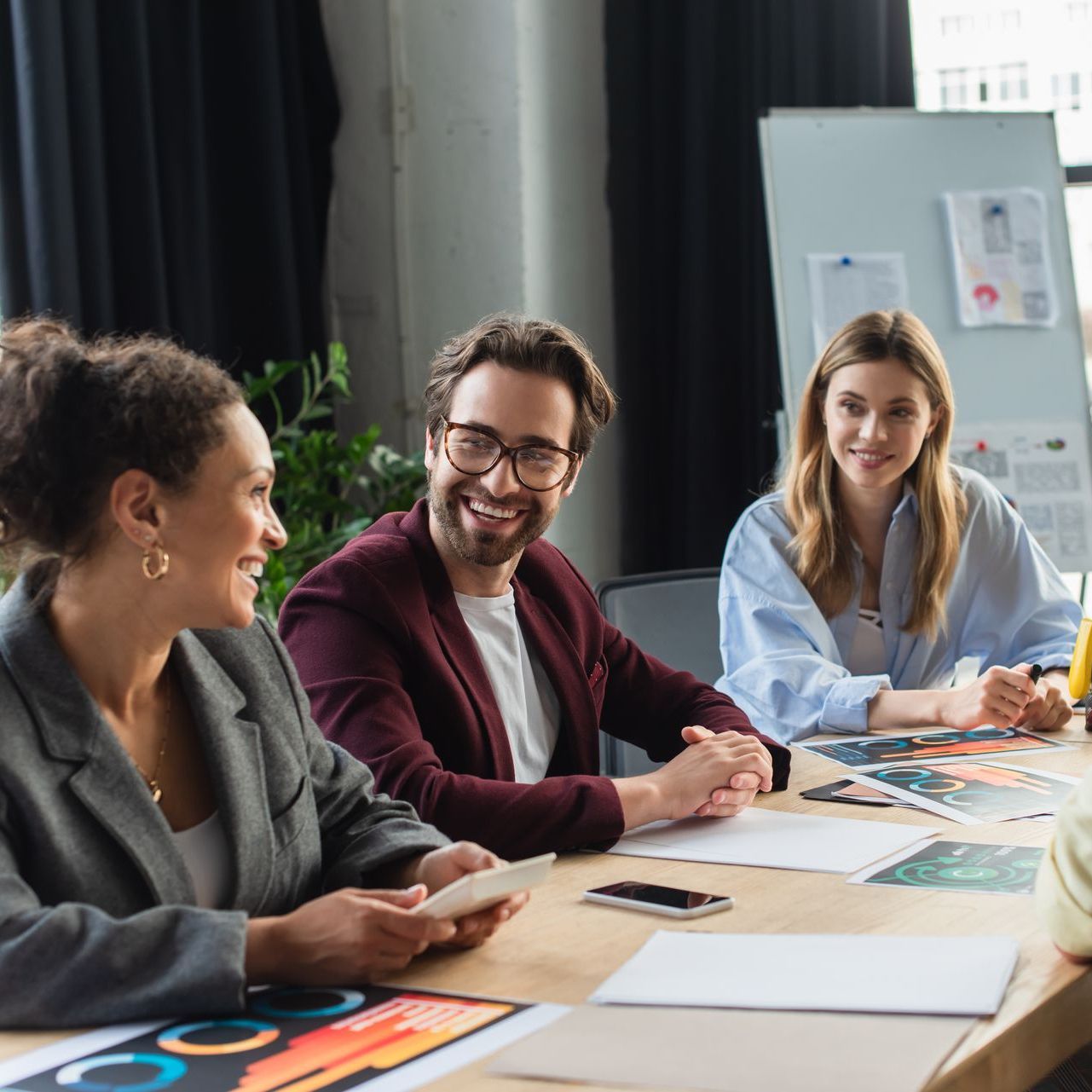 People in a meeting at a table, smiling. A whiteboard and windows are visible in the background.