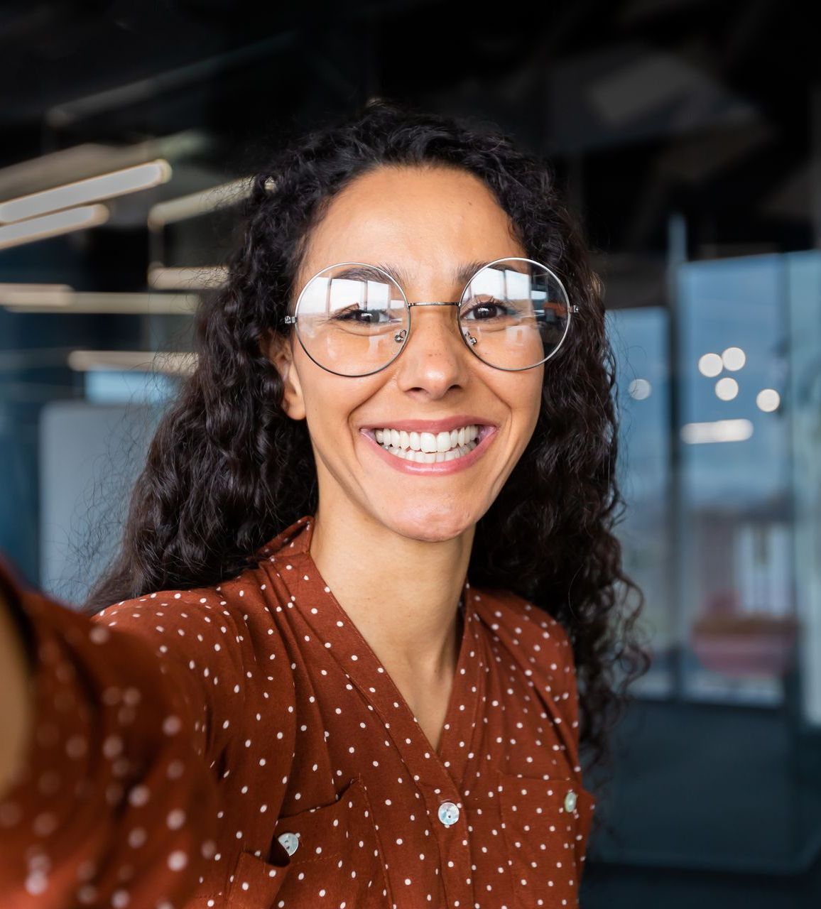 Woman with curly hair and glasses smiles broadly, taking a selfie in an office setting.