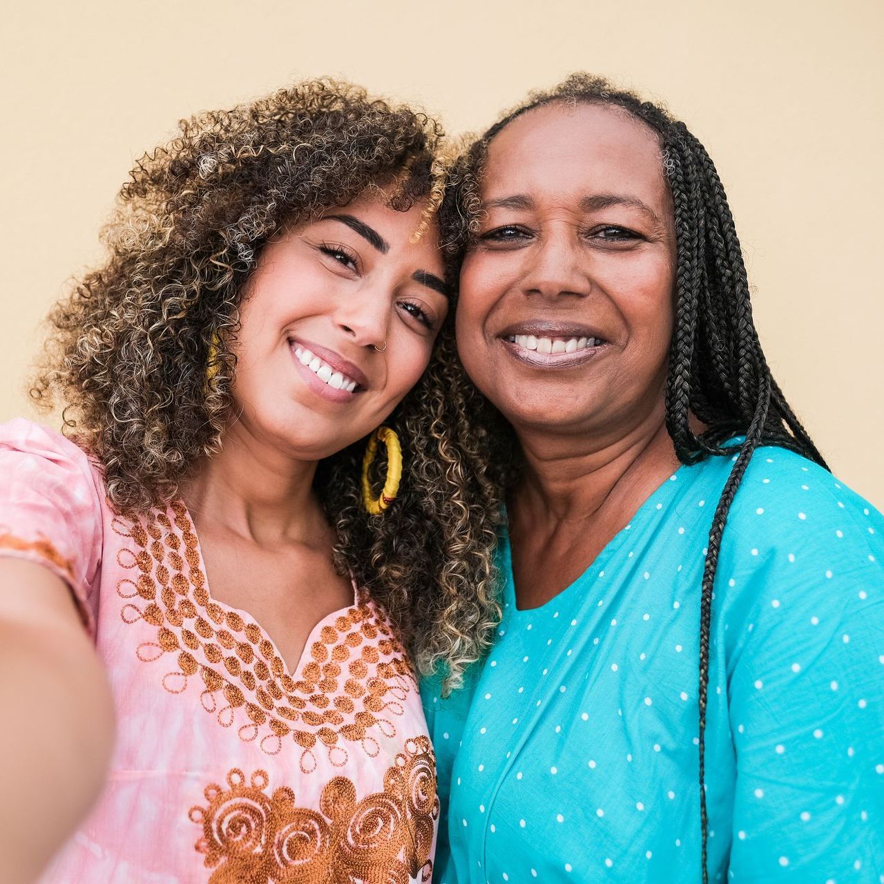 Two women smiling close-up, light-skinned with curly hair and dark braids; pink and blue tops, yellow background.