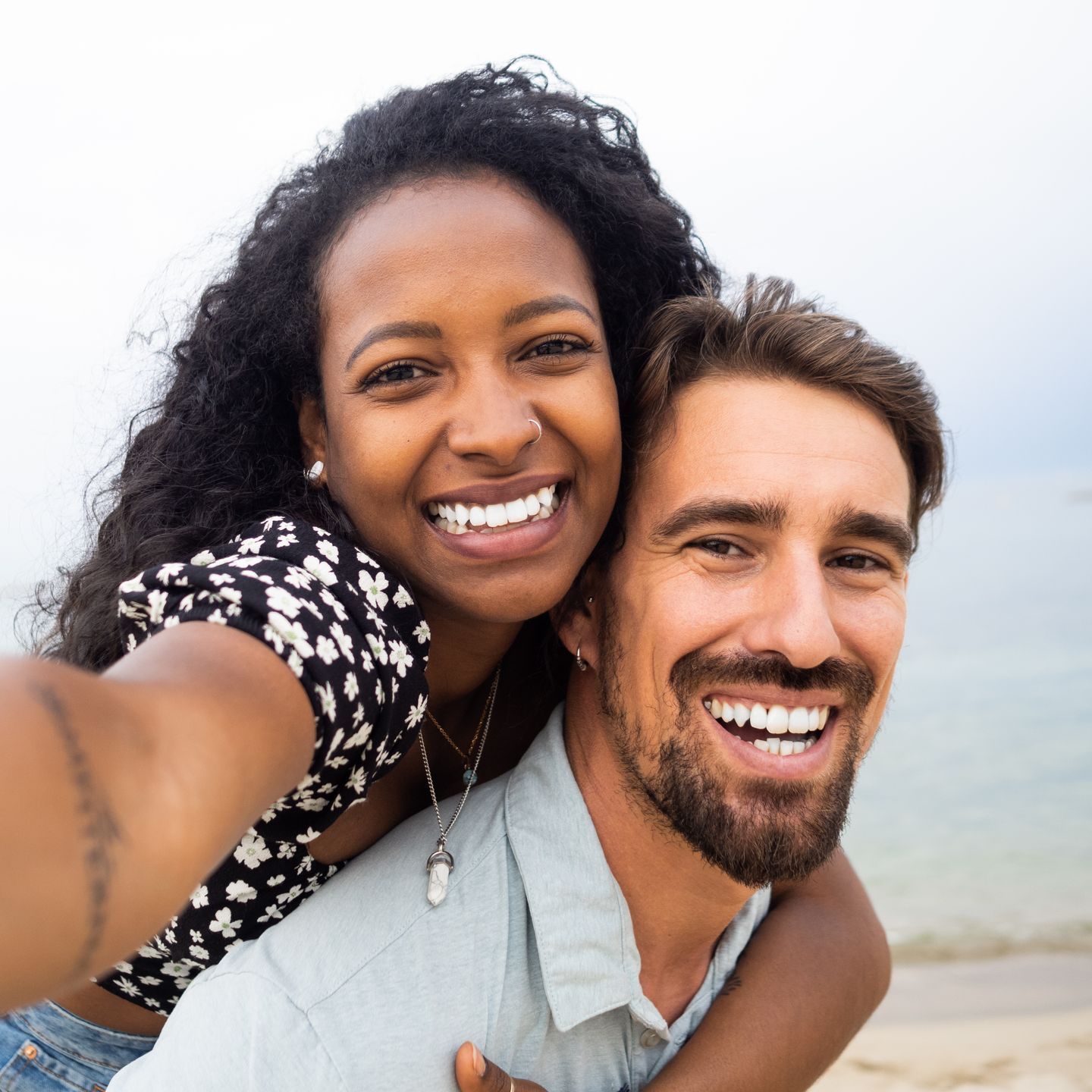 Couple smiling, taking a selfie together at the beach. Woman is on man's back.