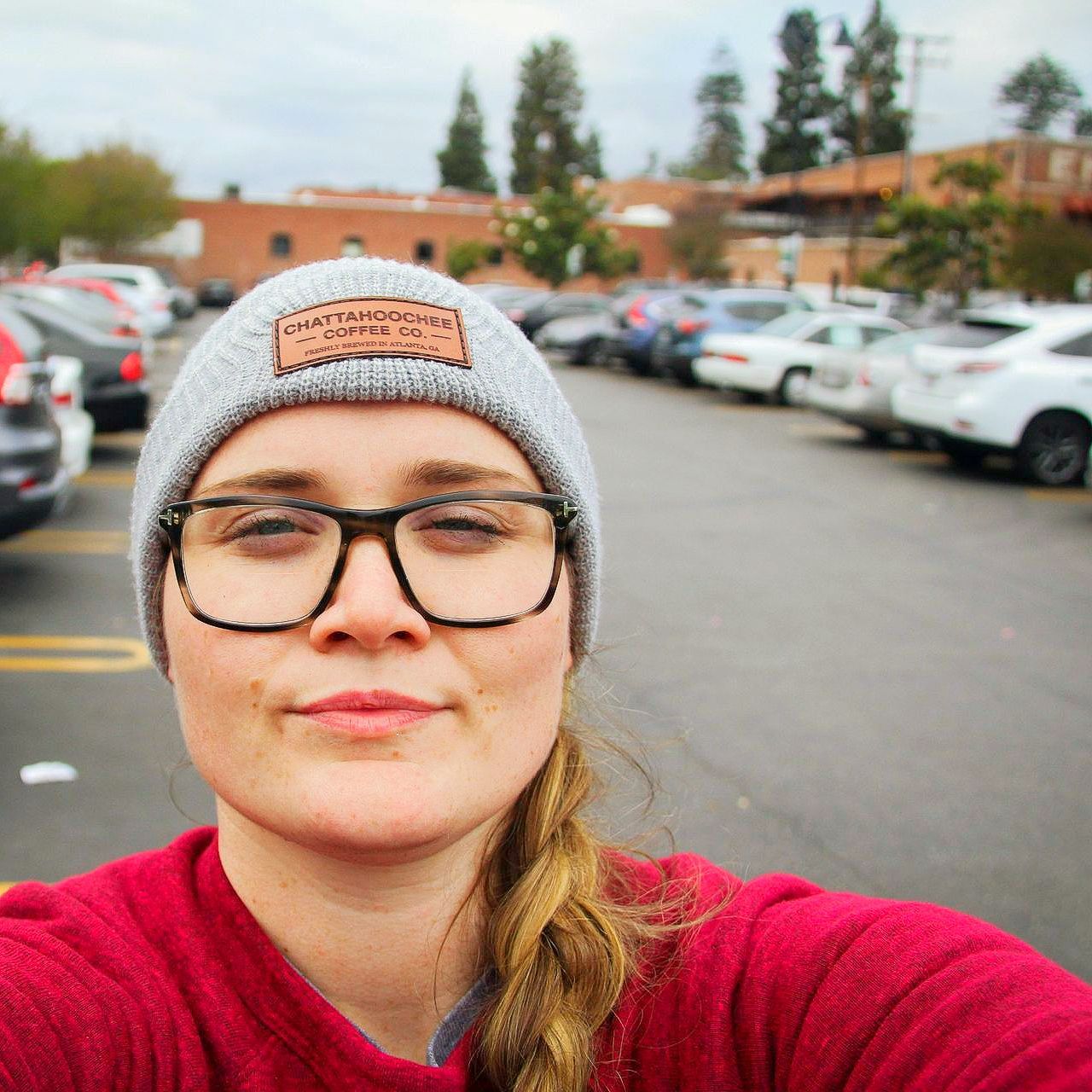 Woman in glasses and beanie smiles in a parking lot, rows of cars behind her.