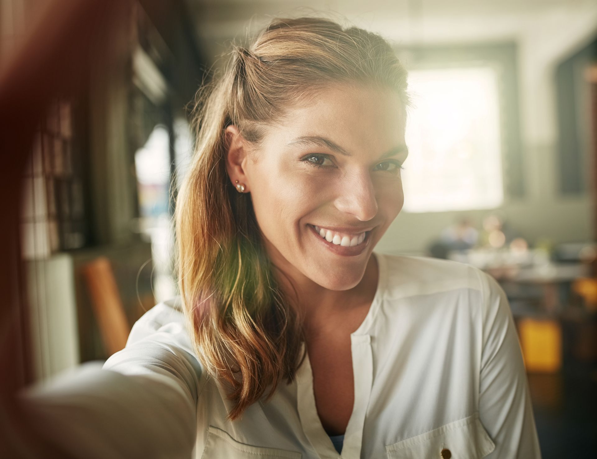 Woman smiling, taking a selfie indoors. She wears a white shirt, with hair half-up. Sunlight in background.