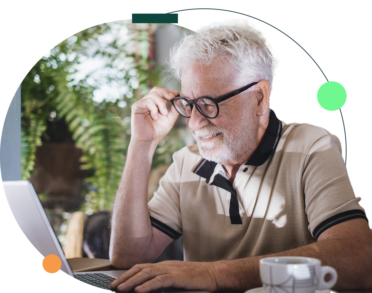 Senior man with glasses using a laptop, smiling, at a cafe table with a coffee cup.