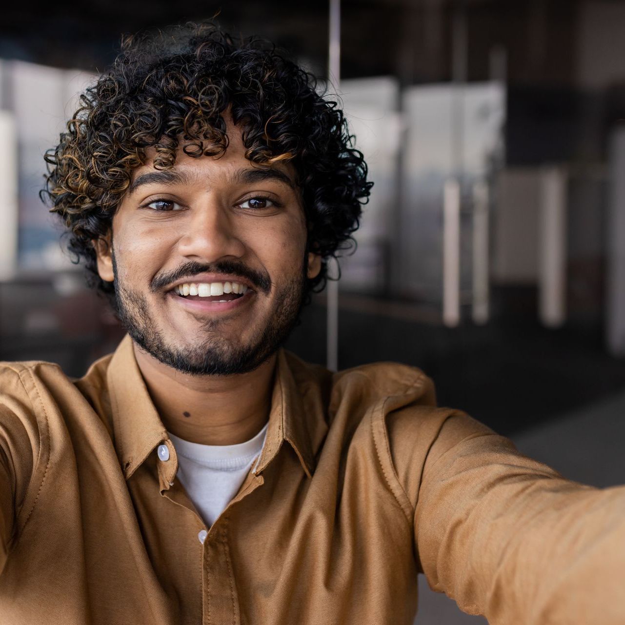 Man with curly hair smiles, taking a selfie in an office setting, wearing a brown shirt.